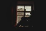 A pensive musician with guitar silhouetted by a window indoors, exuding a moody atmosphere.
