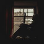 A pensive musician with guitar silhouetted by a window indoors, exuding a moody atmosphere.