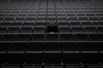 A lone individual sits among rows of empty theater seats, creating a moody atmosphere.