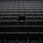 A lone individual sits among rows of empty theater seats, creating a moody atmosphere.