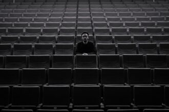 A lone individual sits among rows of empty theater seats, creating a moody atmosphere.