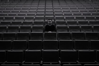 A lone individual sits among rows of empty theater seats, creating a moody atmosphere.