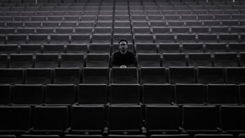 A lone individual sits among rows of empty theater seats, creating a moody atmosphere.
