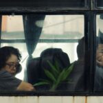 Two people sit inside a bus, viewed through a window, exuding a contemplative mood.